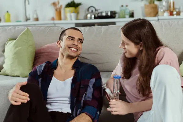 Couple in casual attire enjoy a peaceful moment together on a cozy couch at home, holding glass bong — Stock Photo