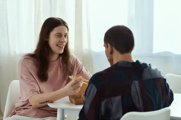 A gay couple in casual clothes enjoying a meal together at a table, sharing food and quality time in their home. — Stock Photo