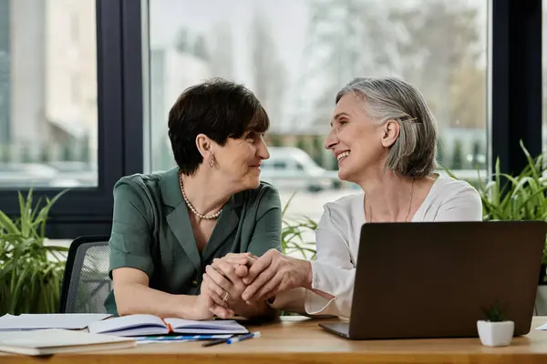 Two mature women are seated at a table, engrossed in work on a laptop. — Stock Photo