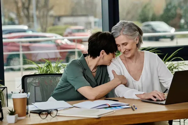 Dos mujeres, una pareja lesbiana madura y hermosa, se sientan en una mesa con un portátil, trabajando juntas en un entorno de oficina. - foto de stock