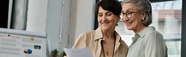 Two mature women, one with dark hair and the other with blonde hair, stand side by side in an office setting. — Stock Photo