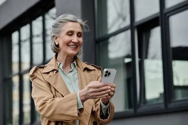 A stylish woman in a trench coat confidently holds a cell phone. — Stock Photo