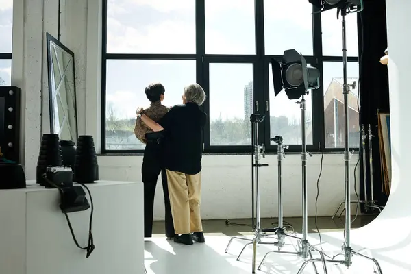 Um casal de lésbicas de meia-idade fica na frente de uma janela, atrás do processo de câmera — Fotografia de Stock