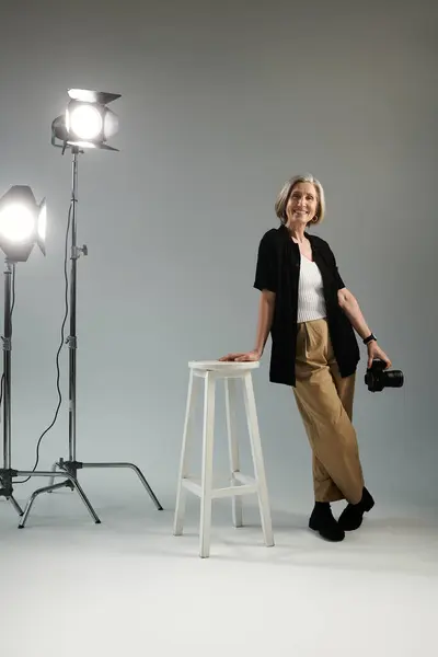 A middle-aged woman stands beside a stool in front of a bright studio light, posing with camera — Stock Photo