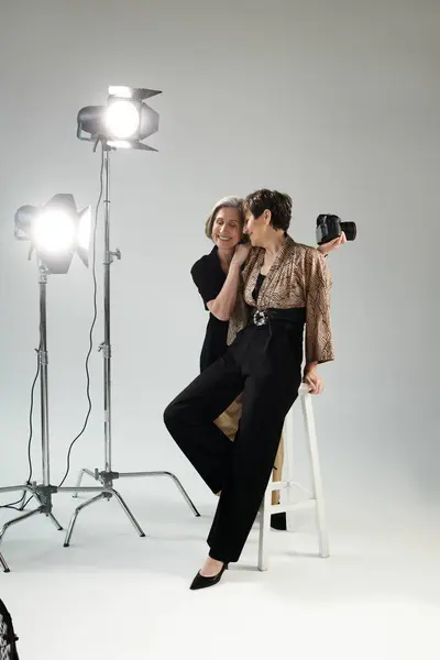 Middle-aged lesbian couple, one as photographer with camera, other as model, sit on stool in photo studio. — Stock Photo