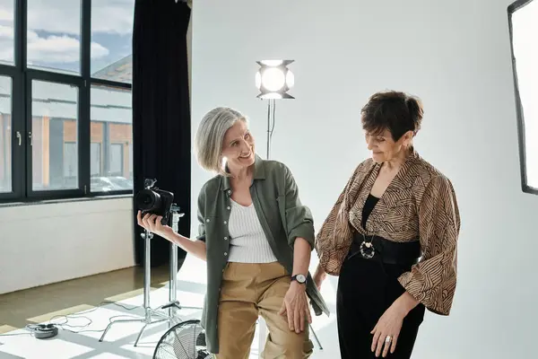 A middle-aged lesbian couple in a photo studio, one holding a camera and the other posing in studio — Stock Photo