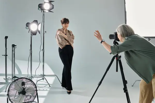 Una pareja de lesbianas de mediana edad en un estudio fotográfico. Una mujer, una fotógrafa, captura a la otra, una modelo, delante de un fan. - foto de stock