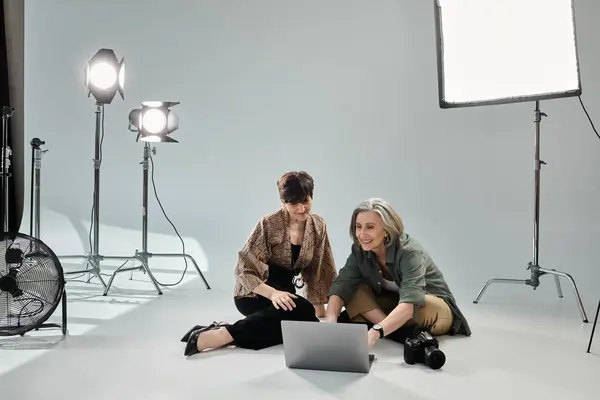 Two middle-aged women sit on floor, engrossed in work on laptop in a photo studio — Stock Photo