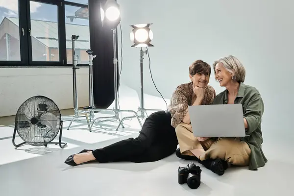 Two middle-aged women, a photographer and model, sitting on the floor together, working collaboratively on a laptop. — Stock Photo