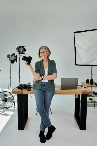 Eine Frau mittleren Alters steht in einem Fotostudio und hält eine Kamera vor einen Schreibtisch. — Stockfoto