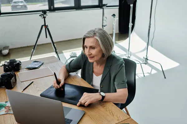 Frau mittleren Alters mit Laptop und Grafik-Tablet am Tisch in modernem Fotostudio. — Stockfoto