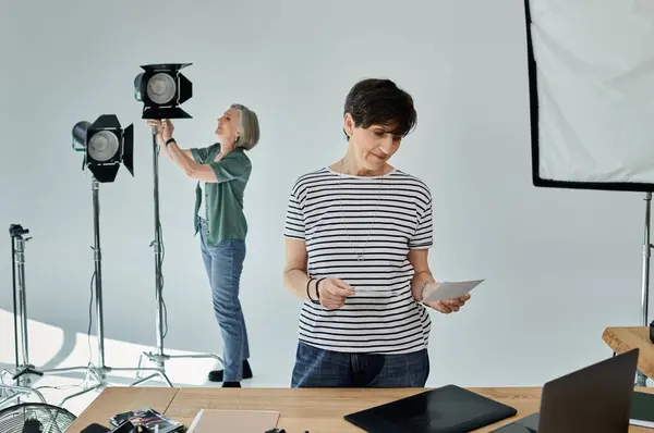 Una mujer de mediana edad y su colega están lado a lado en un moderno estudio fotográfico, listos para trabajar juntos en un proyecto. - foto de stock