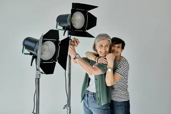 A middle-aged lesbian couple standing and hugging in front of bright lights in a modern photo studio. — Stock Photo