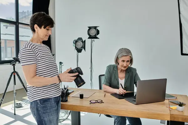 A middle-aged woman sits at a table with laptop, other one with a camera in front of her, ready to capture moments in a professional setting. — Stock Photo