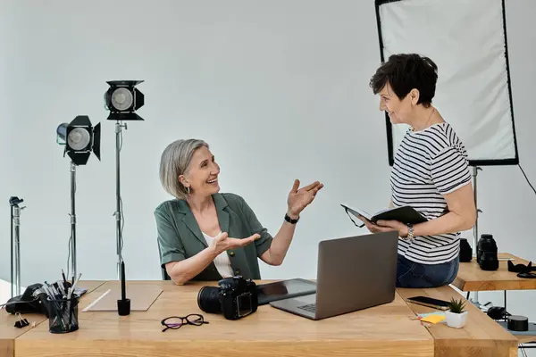 Two middle-aged women engage in dialogue while working in front of a laptop in a modern photo studio. — Stock Photo