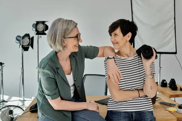 Two middle aged women sit on a table, engaging with the camera in a professional studio setting. — Stock Photo