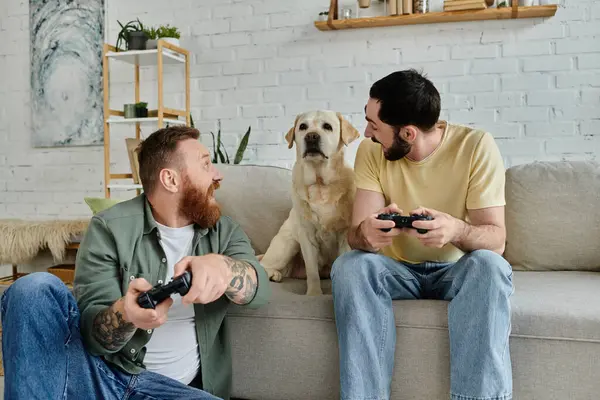 Dois homens, desfrutando de uma sessão de jogo em um sofá com foco intenso e emoção, enquanto seu labrador leal observa de perto. — Fotografia de Stock