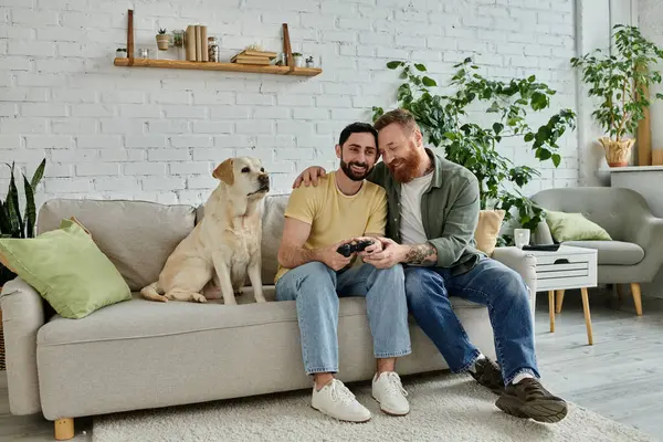 Due uomini, una coppia gay, si siedono su un divano a giocare al videogioco con il loro cane labrador in un ambiente accogliente soggiorno. — Foto stock