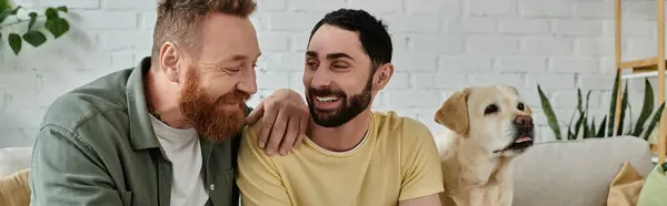 A bearded gay couple sit on a couch with their Labrador dog nearby in their cozy living room. — Stock Photo