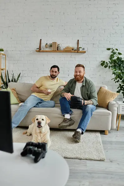 Two bearded men relax on a couch with their Labrador dog, watching a baseball match on TV in a cozy living room. — Stock Photo
