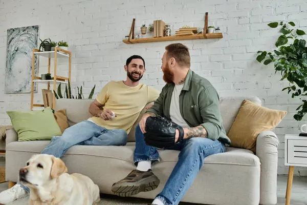 Due uomini barbuti si siedono su un divano, guardando una partita di sport con il loro cane labrador al loro fianco. — Foto stock