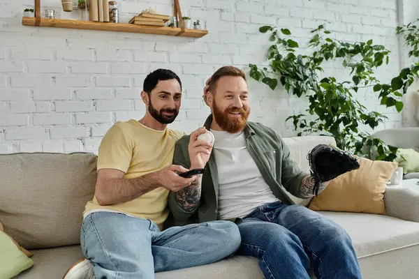 Um duo de homens, casal gay barbudo, se senta no topo de um sofá enquanto assiste a um jogo esportivo — Fotografia de Stock