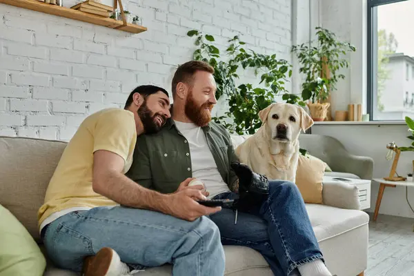 Dois homens, um casal gay, sentam-se em um sofá assistindo a um jogo esportivo com seu cão Labrador em um ambiente acolhedor sala de estar. — Fotografia de Stock