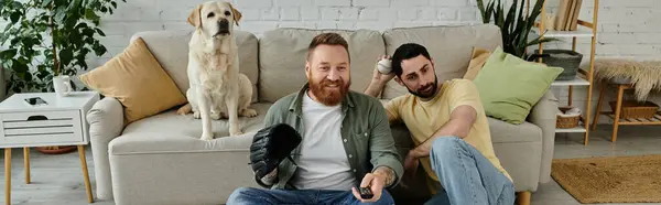 Dois homens com barbas se sentando em um sofá, absorvidos em assistir a um jogo esportivo com seu cão Labrador ao seu lado. — Fotografia de Stock