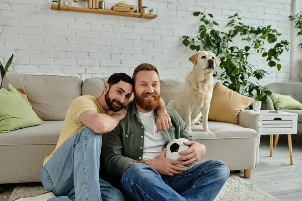 Dois homens com barba sentam-se num sofá, a ver um jogo desportivo, com um cão Labrador ao fundo. — Fotografia de Stock