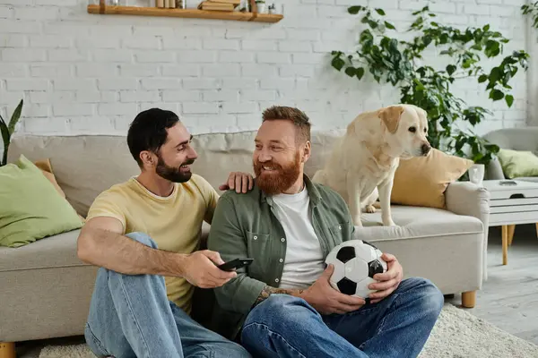 Two men with a soccer ball, lounging on a couch with their labrador dog, engrossed in a sports match on TV. — Stock Photo