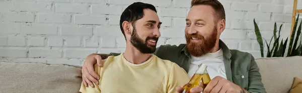 Bearded gay couple clinking beer bottle and sitting on couch in living room. — Stock Photo