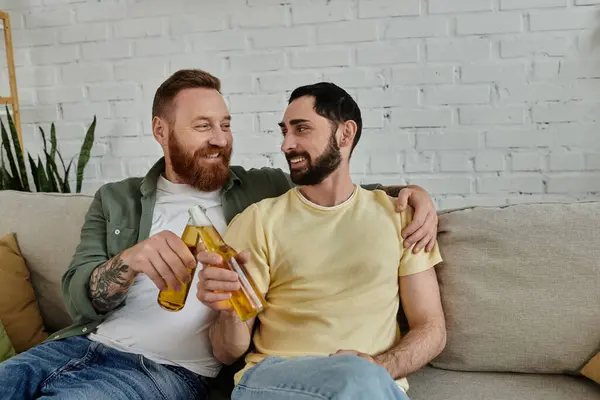 Barbudo feliz gay casal relaxa no sofá clinking cerveja garrafas no casa — Fotografia de Stock