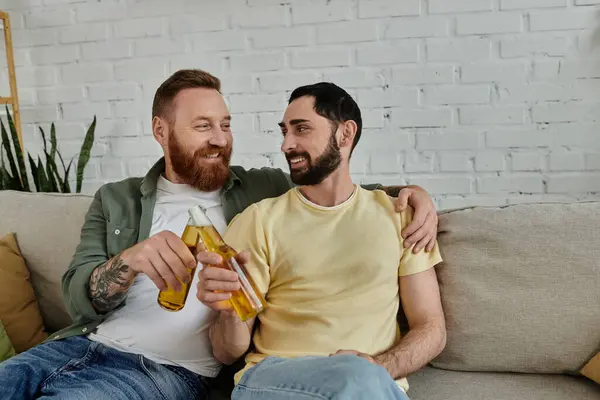 Bearded happy gay couple relaxes on sofa clinking beer bottles at home — Stock Photo