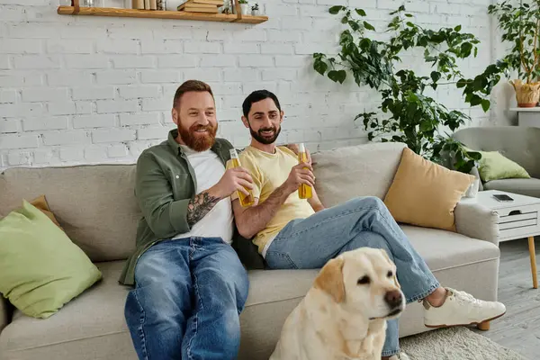 Two men, a gay couple, sit on a couch with their dog, watching a sports match together in their living room. — Stock Photo
