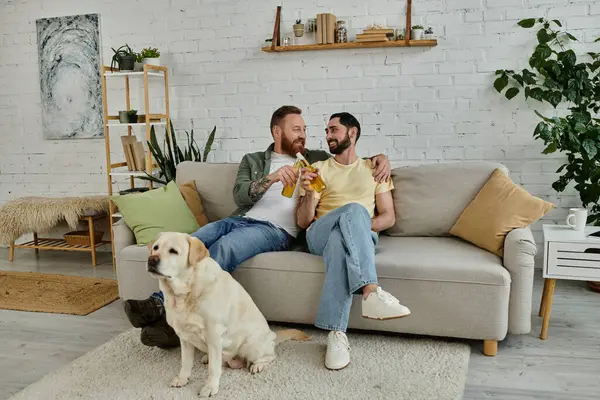 Dois homens, um casal gay barbudo, sentam-se em um sofá assistindo a um jogo esportivo com seu cão labrador ao seu lado na sala de estar. — Fotografia de Stock