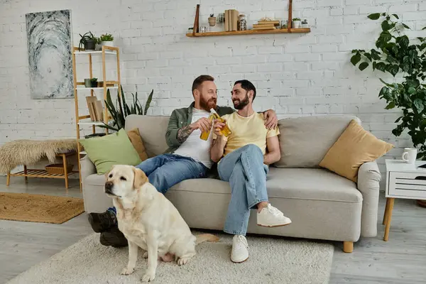 Dois homens, um casal gay barbudo, sentam-se em um sofá assistindo a um jogo esportivo com seu cão labrador ao seu lado na sala de estar. — Fotografia de Stock