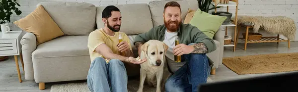 Two men, a bearded gay couple, sit on a couch with their labrador dog in a living room, engrossed in watching a sports match. — Stock Photo