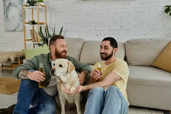 Dois homens barbudos relaxam em um sofá com seu labrador, assistindo a um jogo esportivo em sua sala de estar. — Fotografia de Stock