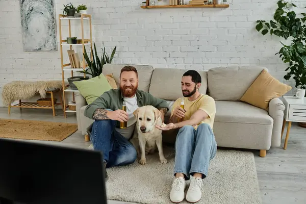 Two bearded men sit on a couch with their Labrador, enjoying a sport match together in their living room. — Stock Photo