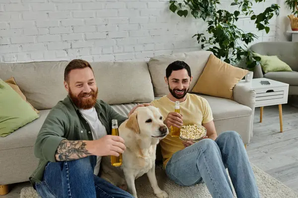 Dois homens barbudos alegremente assistir a um jogo de esportes com seu cão labrador no sofá na sala de estar. — Fotografia de Stock