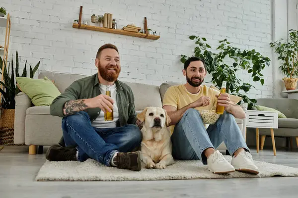 Two men with beards relax on a couch, watching a sports match with their Labrador dog in the living room. — Stock Photo