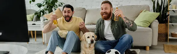 Um casal gay e seu Labrador sentados em cima de um sofá, assistindo a um jogo de esportes na sala de estar. — Fotografia de Stock