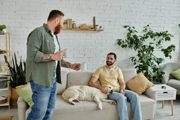 Two men, one standing with cups, one sitting on a couch, in a living room with a labrador dog nearby. — Stock Photo