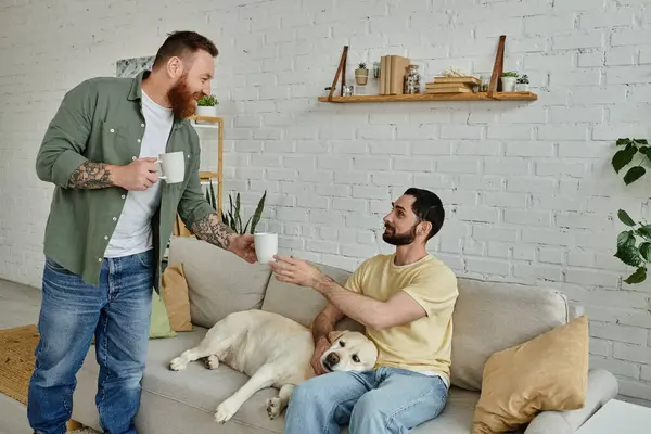Bearded man sitting on a couch next to a labrador dog, his partner offering drink — Stock Photo