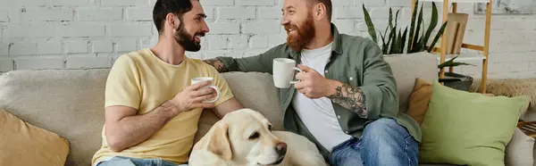 Bearded gay couple sits on couch with Labrador dog while holding cups of coffee in cozy living room. — Stock Photo