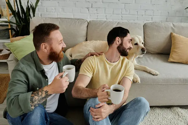 Двоє чоловіків, бородата гей-пара, тримають чашки кави біля собаки Лабрадору у своїй вітальні. — стокове фото
