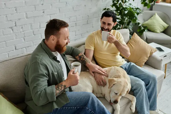 Two men, both with beards, drinking coffee in their living room while sitting on a couch with their beloved labrador. — Stock Photo