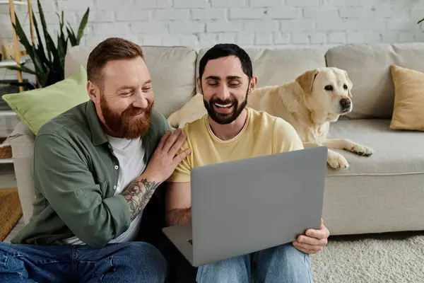 Bearded gay couple on a couch, working on a laptop, alongside a labrador dog in a cozy living room setting. — Stock Photo