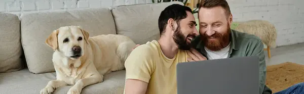 Casal gay barbudo e seu cão Labrador sentado em um sofá, ambos absorvidos na visualização de conteúdo em um laptop. — Fotografia de Stock