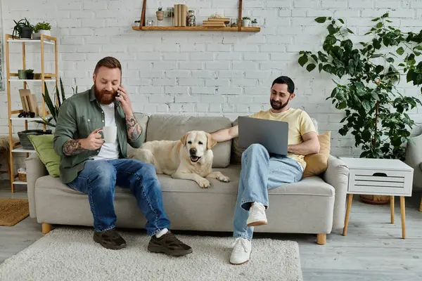 Um casal gay barbudo está trabalhando remotamente em um laptop enquanto está sentado em um sofá com seu cão labrador em uma acolhedora sala de estar. — Fotografia de Stock